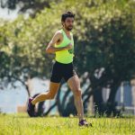 Full length of young bearded sportsman in activewear running on grassy lawn in city park near green trees during outdoor workout