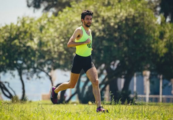 Full length of young bearded sportsman in activewear running on grassy lawn in city park near green trees during outdoor workout