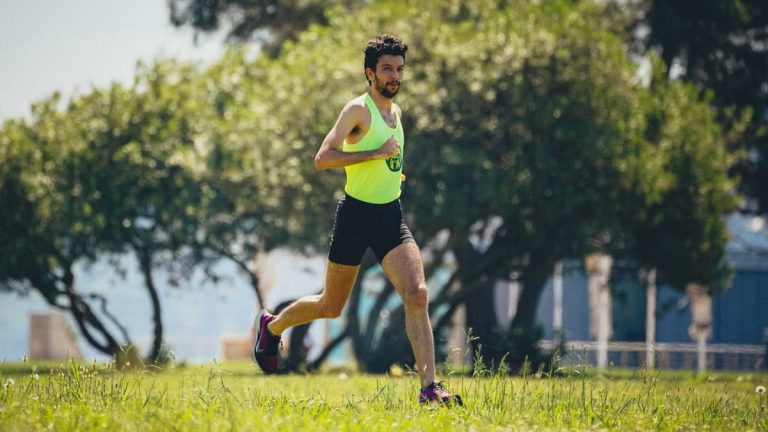 Full length of young bearded sportsman in activewear running on grassy lawn in city park near green trees during outdoor workout